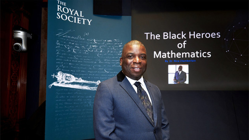 Nira stands infront of a Royal Society board and a presentation titled' The Black Heroes of Mathematics'.